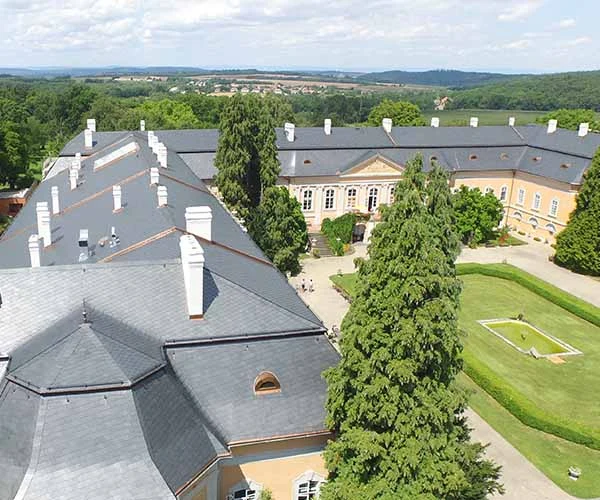 Velká budova s břidlicovou střechou na pozadí zelené krajiny.