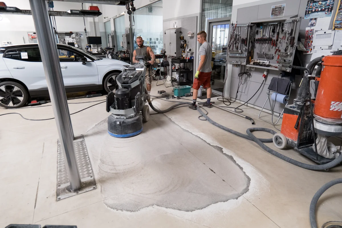 Muži brousí betonovou podlahu v dílně s průmyslovým zařízením.