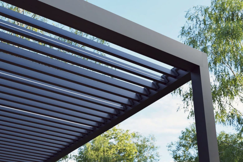 Moderní bioklimatická pergola s nastavitelnými lamelami na pozadí zelených stromů.