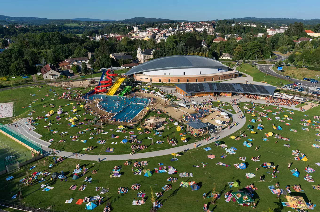 Pohled na venkovní koupaliště s bazénem, skluzavkami a slunečníky.