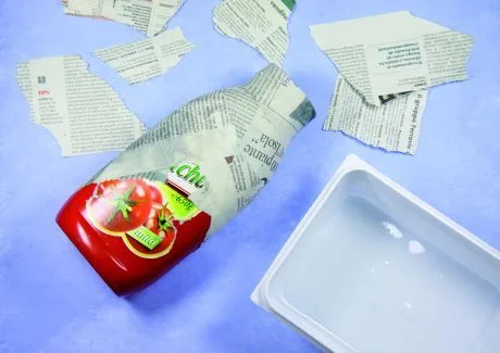 Ketchups v novinách vedle plastového kontejneru na modrém pozadí.
