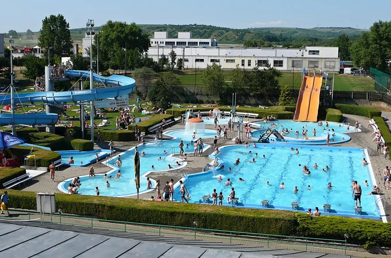 Venkovní aquapark s bazénymi a vodními skluzavkami plný lidí.