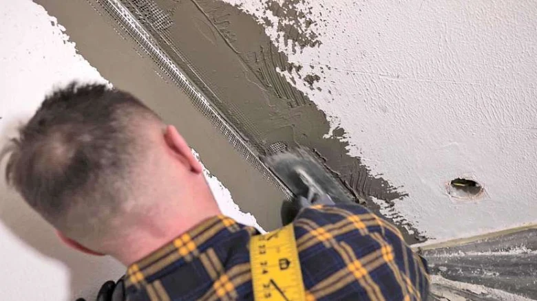 Muž lepi síťovinu na rohu stěny špachtlí s hmotou.
