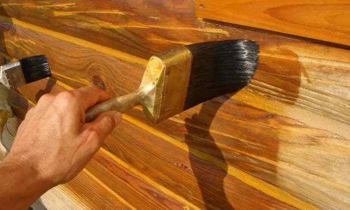 Ruka nanáší venkovní lazuru na dřevěné prkno štětcem.