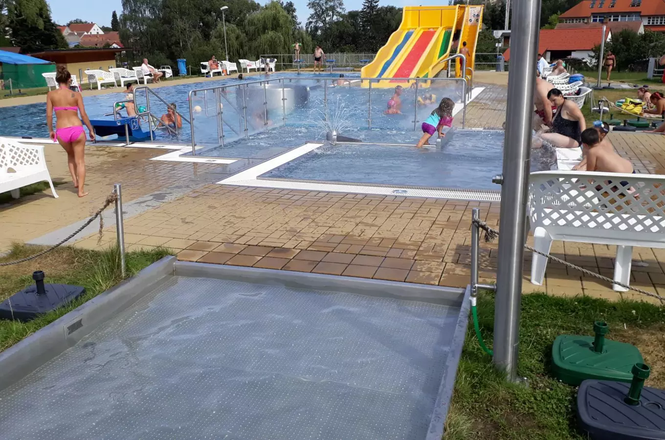 Venkovní bazén ve Velešíně s vodním skluzavkou a dětmi hrajícími si v koupališti.