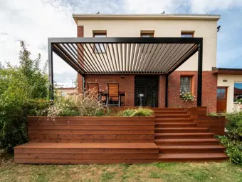 Bioklimatická pergola s dřevěnou terasou vedoucí k vchodu do domu.