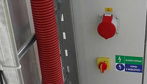 Hlavní vypínač v rozvaděči s varovnými štítky a červeným kabelovým potrubím.