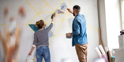 Muž a žena malují žluté geometrické vzory na bílou stěnu.