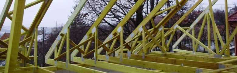 Žluté dřevěné vazníky tvořící střešní konstrukci na pozadí venkovního prostředí.
