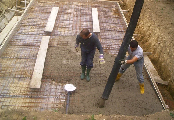 Dva dělníci nalévají beton na základnu bazénu s výztuží.