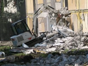 Bílý bagr rozebírá žlutou budovu na místě demolice.