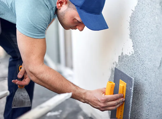 Muž opravuje venkovní fasádu aplikací omítky na betonovou stěnu.