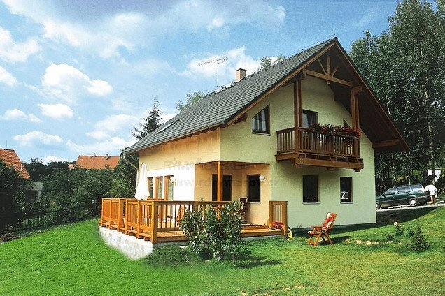 Dřevostavba s terasou na pozadí zelené trávy pod modrou oblohou.