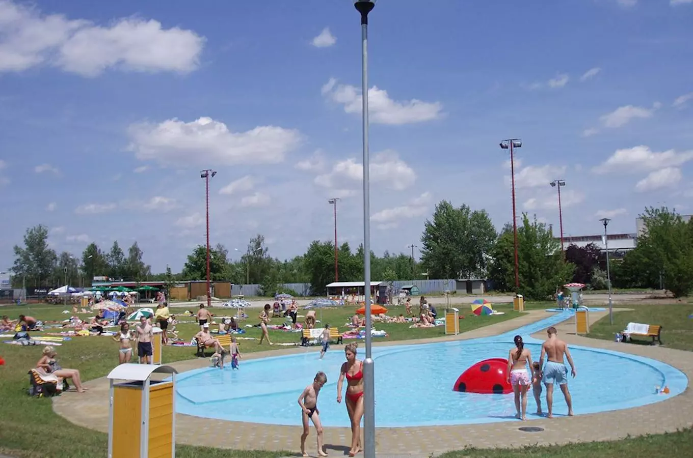 Venkovní bazén v Chropyni s lidmi plavajicimi si užívajících vodních atrakci pod sluncem.