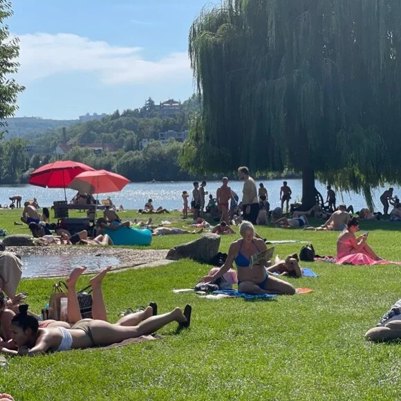 Lidé se opalují na trávníku u jezera v Praze.