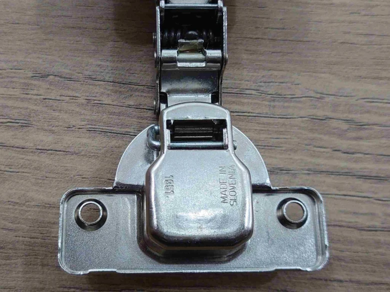 Stříbrný pant pro skříně s viditelným pružinovým mechanismem.