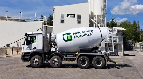 Bílý betonový míchač značky Heidelberg Materials u průmyslové budovy.