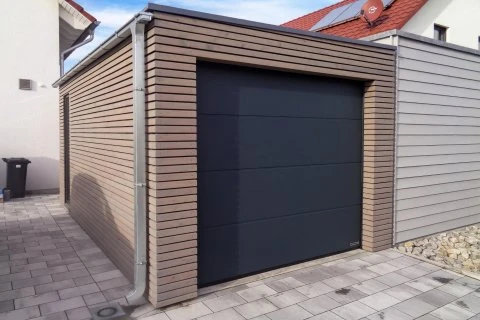 Moderní dřevěná garáž s tmavým posuvným vratem na dlažbě.