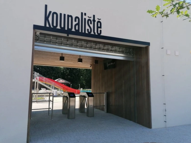 Vstup do koupaliště s automatickými brankami a vodním skluzem na pozadí.